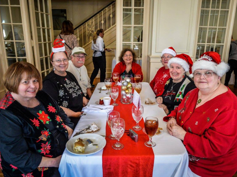 Seven people in Christmas attire seated around a rectangle table at Festival of Trees luncheon.